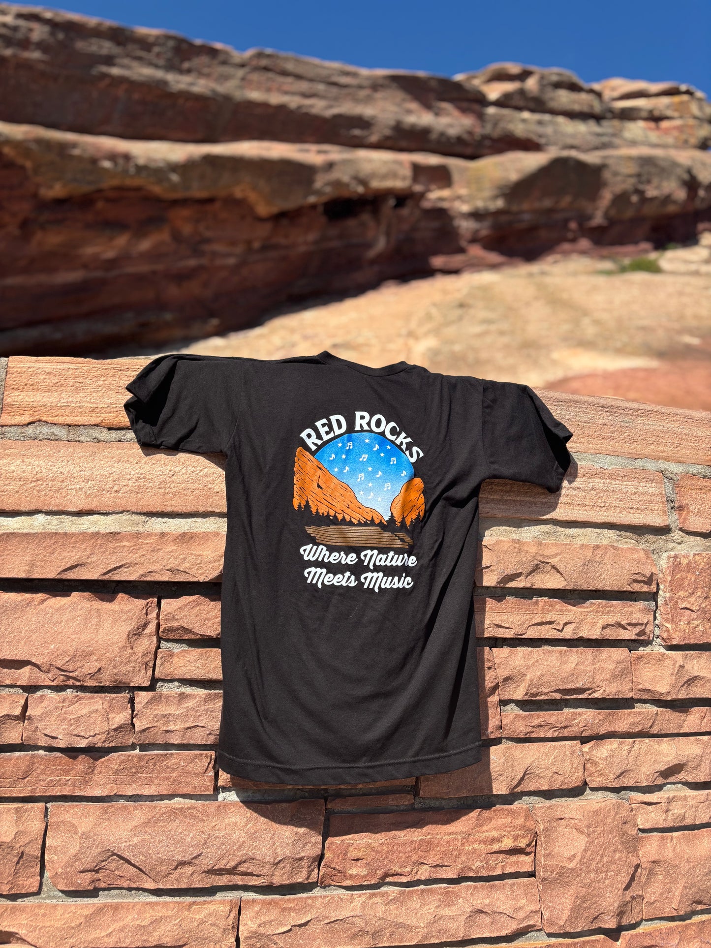 Black t-shirt with Red Rocks Amphitheatre graphic on a stone wall with red rock formations in the background.